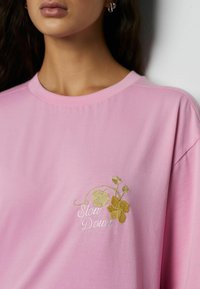Lyserød bomulds-T-shirt med broderet gulddetalje af blomster og teksten "Slow Down" på venstre bryst. Blød, afslappet pasform og rund halsudskæring.