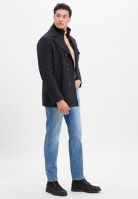 Mann in einem dunkelblauen Peacoat, beigem Pullover, hellblauen Jeans und schwarzen Stiefeln, der mit einer Hand in der Tasche seitlich schaut.