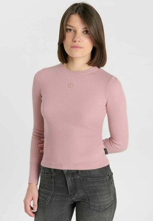 Junge Frau mit schulterlangen braunen Haaren, die ein rosa geripptes Langarmshirt und dunkelgraue Jeans trägt, vor einem schlichten Hintergrund stehend.