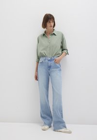 Grüne knopfbare Hemd mit subtilem Punktmuster, kombiniert mit hellblauen weit geschnittenen Jeans. Das Model trägt beige Schuhe vor einem weißen Hintergrund.