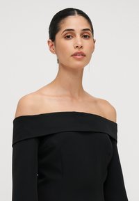 Svart avskjæringskjole med lange ermer i en tettsittende design. Glatt stoff med strukturert utringning og minimale dekorasjoner.