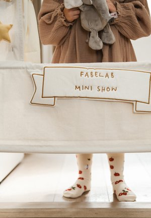 Bavlněný herní stan s krémovou látkou, zlatou výšivkou s textem „FABELAB MINI SHOW“ a dítětem v ponožkách s vzorem třešní, které drží šedou hračku slona.