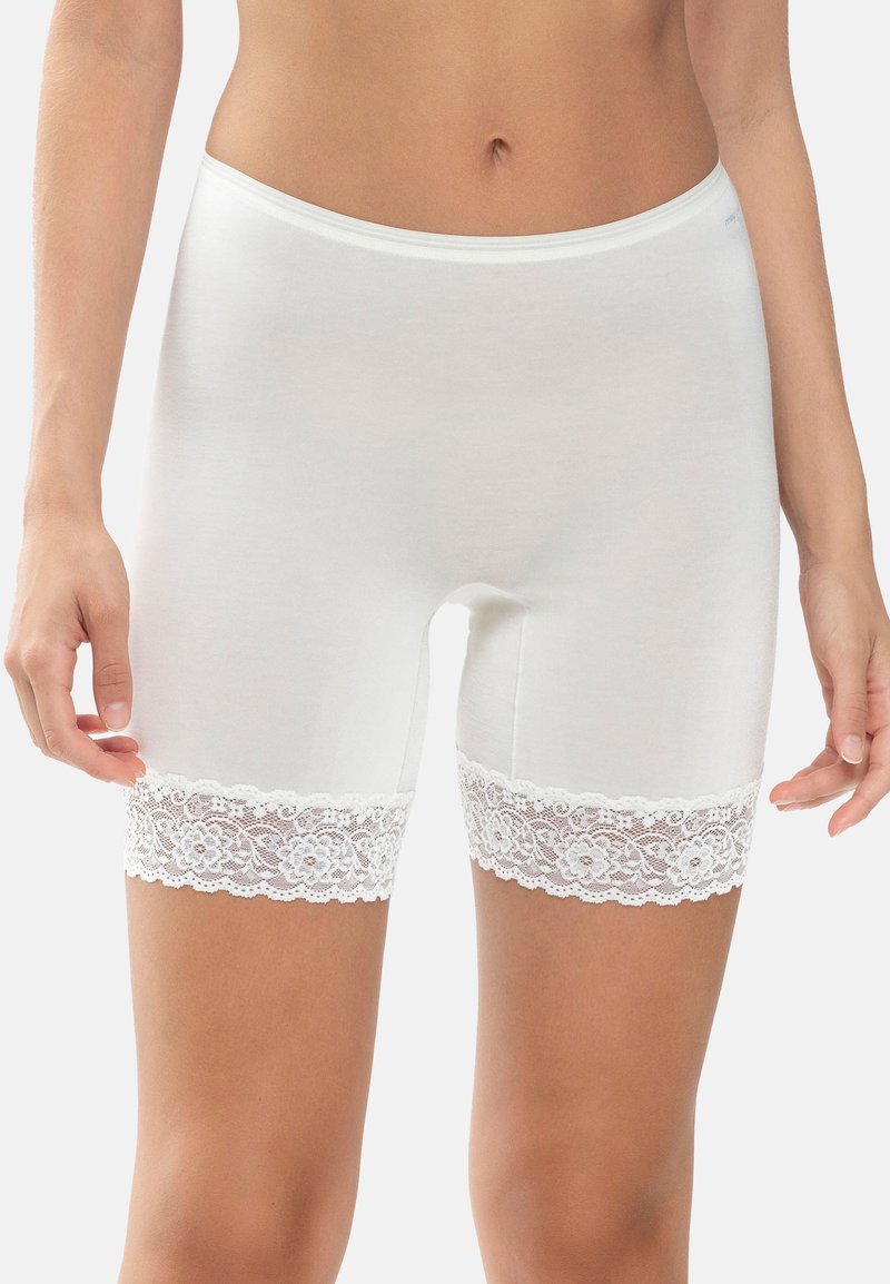 Donna che indossa biancheria intima bianca fino alla metà della coscia con bordo in pizzo floreale, in piedi con le mani rilassate ai lati.