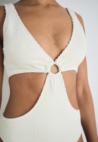 Maillot de bain blanc texturé une pièce avec un décolleté plongeant, une découpe circulaire au centre et des bretelles larges pour un soutien.