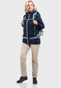 Marineblaue wasserdichte Jacke mit weißem Reißverschluss, beigen Hosen und schwarzen Wanderschuhen. Ausgestattet mit einem hellblauen Rucksack für Outdoor-Aktivitäten.