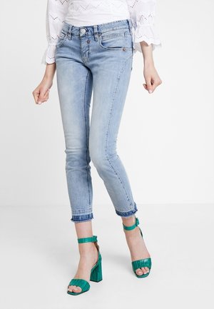 Frau trägt hellblaue, kurz geschnittene Skinny-Jeans mit ausgefransten Säumen und grüne High-Heel-Sandalen, steht vor weißem Hintergrund.