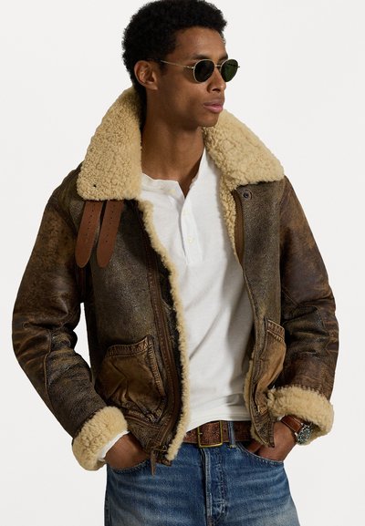 Καφέ δερμάτινο μπουφάν bomber με ταμπά γούνινο γιακά και μανίκια, που διαθέτει μεταλλικά αξεσουάρ και καφέ δερμάτινους ιμάντες. Κάτω από αυτό, ένα κανονικό λευκό πουκάμισο.
