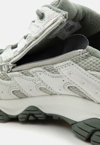 Baskets grises et blanches avec fermeture éclair latérale partiellement ouverte, montrant un col rembourré et les détails de la semelle texturée en gros plan.
