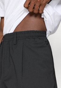 Melni, teksturēti bikses ar elastīgu jostasvietu, smalkām krokām un nelielu logotipa akcentu sānos. Apvienots ar baltu krekli.