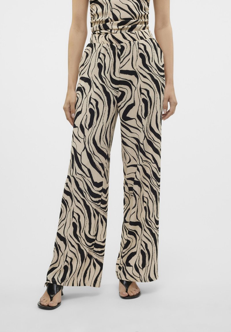Brede broek in een beige stof met een zwart golvend patroon, voorzien van een tailleband en zijzakken, gedragen met zwarte sandalen.