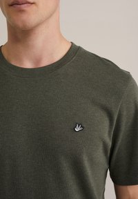 Camiseta de cuello redondo verde oliva hecha de telas suaves, con un pequeño logotipo de pájaro bordado en el lado izquierdo del pecho. Diseño simple y casual.