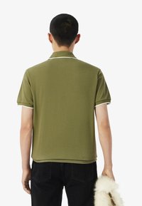 Polo in cotone verde oliva con maniche corte, rifinitura del colletto bianco e orlo dritto, indossato con pantaloni neri e un accessorio in pelliccia.