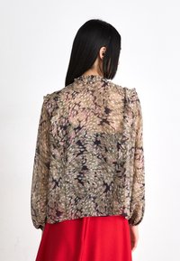 Blusa floreale trasparente in toni smorzati di verde, rosa e nero, con spalle arricciate e polsini elasticizzati, indossata con una gonna rossa.