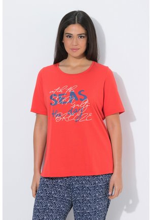 Frau mit langen dunklen Haaren, die ein rotes T-Shirt mit Grafik und marineblaue gemusterte Hose trägt, vor einem weißen Hintergrund stehend.