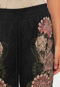 Black pants with elastic waistband and drawstring, featuring intricate pink and beige floral embroidery on the thigh, worn with beige knitted top.