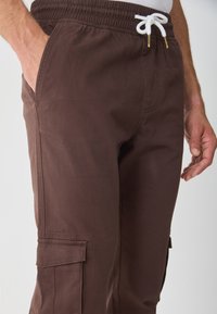 Hnědé cargo kalhoty s elastickým pasem a šňůrkou. Mají boční kapsy a hladkou texturu. Navrženy pro volnočasové nošení.