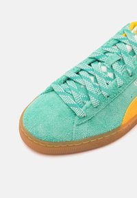 Green suede sneaker with textured finish, patterned laces, gum rubber sole, and yellow accent on the side. Close-up view of the toe area.