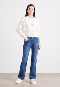 En kvinne står selvsikkert, iført en kremstripet cardigan, blåjeans og hvite sneakers, med en nøytral bakgrunn som fremhever antrekket hennes.