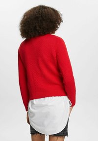 Pull rouge en tricot avec un motif texturé en losanges, col et ourlet côtelés. Porté par-dessus une chemise blanche, avec un dos arrondi et plus long.