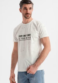 Camiseta de algodón blanca con una ligera textura, que presenta una impresión negra: "TU ÚNICO LÍMITE ERES TÚ MISMO". Corte casual, mangas cortas.