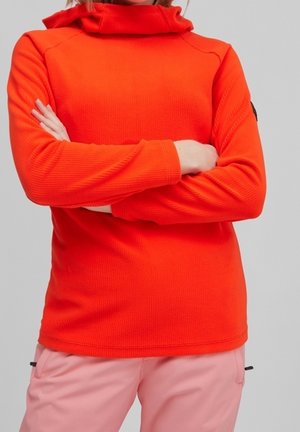 Person mit verschränkten Armen, die ein helloranges Langarmshirt und eine hellrosa Hose trägt, steht vor einem schlichten hellgrauen Hintergrund.