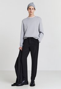 Uomo che indossa un berretto grigio, un pullover grigio, pantaloni neri e scarpe nere, mentre tiene una giacca nera nella mano sinistra.