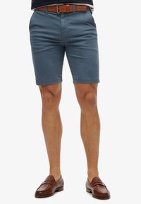 Pantaloni corti blu in cotone con fronte piatto, tasche laterali e orli arrotolati, abbinati a mocassini in pelle marrone e una cintura abbinata.