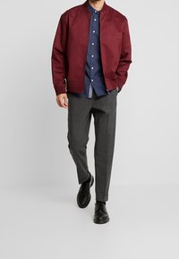 Giacca bomber bordeaux con colletto a stand, abbinata a una camicia blu navy a pois e pantaloni grigi su misura. Le scarpe nere completano l'outfit.
