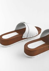Sandales à glissière blanches en matériau synthétique avec une semelle en bois, présentant une sangle supérieure incurvée ornée de clous en argent et un pied qualitatif texturé.