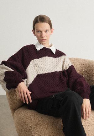 Knit sweater in dark burgundy and cream, featuring a zigzag cable pattern and oversized sleeves. Worn over a white collared shirt.