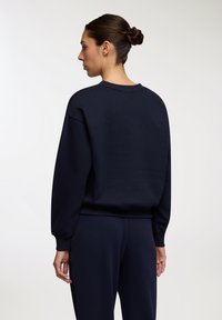 Granatowy sweatshirt o luźnym kroju, z okrągłym dekoltem i ściągaczami, wykonany z miękkiego materiału, zaprezentowany od tyłu z dopasowanymi spodniami.
