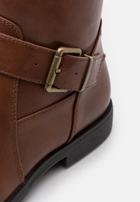 Brązowy skórzany botek za kostkę z przeszywanym wzorem, metalowym zapięciem i czarną gumową podeszwą. Gładka tekstura o matowym wykończeniu.