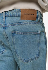 As calças de ganga azul-claro apresentam um patch de couro castanho com "PEGADOR" em relevo. O tecido mostra uma ligeira textura com detalhes costurados.