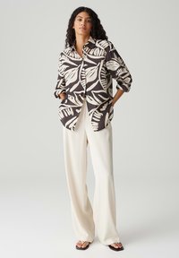 Camicia con bottoni caratterizzata da una grande stampa botanica beige su uno sfondo marrone scuro. Presenta due tasche frontali e maniche arrotolate.