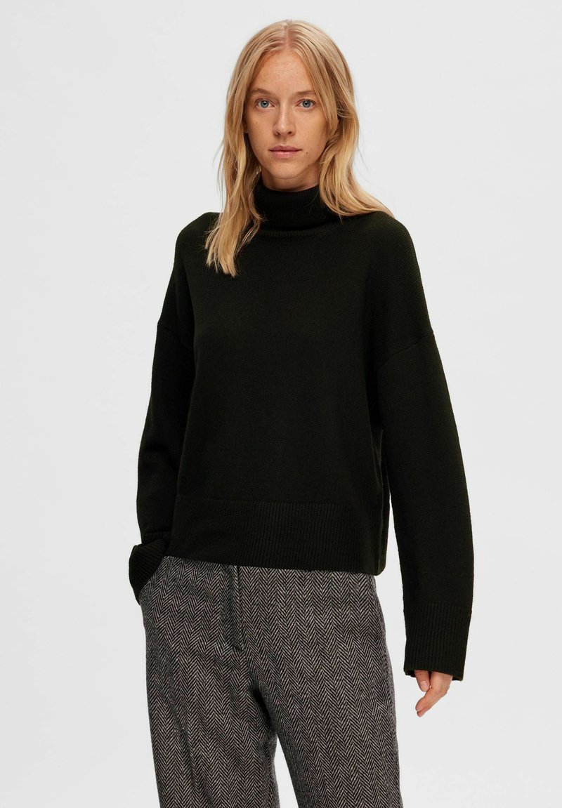 Pull à col roulé vert foncé avec une coupe décontractée, des épaules tombantes et des poignets côtelés. Associé à un pantalon en pied-de-poule grise à motifs.