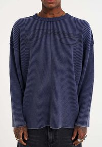 Marineblaues, geripptes Langarmshirt mit lockerer Passform, ausgestattet mit einem Rundhalsausschnitt und einem dezenten Grafikdruck auf der Vorderseite. Weiche Textur und lässiger Stil.