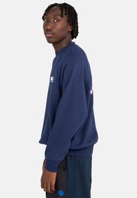 Sudadera azul marino con cuello redondo y puños acanalados, que presenta un pequeño parche de logo en el pecho y un diseño impreso en la parte posterior.