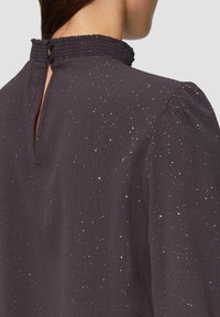 Dunkellila Bluse aus strukturiertem Stoff, mit einem hohen Rüschenausschnitt und einer kleinen Schlüsselöffnung auf der Rückseite, akzentuiert durch Glitzer.