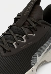 Nike trail hardloopschoen in donkergrijs met een gestructureerde bovenkant, contrasterend wit mesh-paneel en gemêleerde veters; voorzien van "Nike Trail" branding.