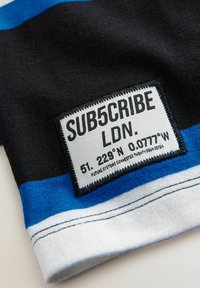 Svart, blå och vit tyg med en rektangulär etikett. Etiketten har texten "SUB5CRIBE LDN" och koordinater, med svarta sömnadsdetaljer.