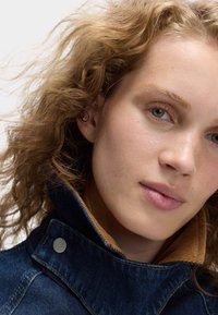 Jeansowa kurtka z wysokim kołnierzem, z kontrastowym podbiciem z korduru. Tkanina jest ciemnoniebieska z widocznymi szwami i metalowymi zatrzaskami.