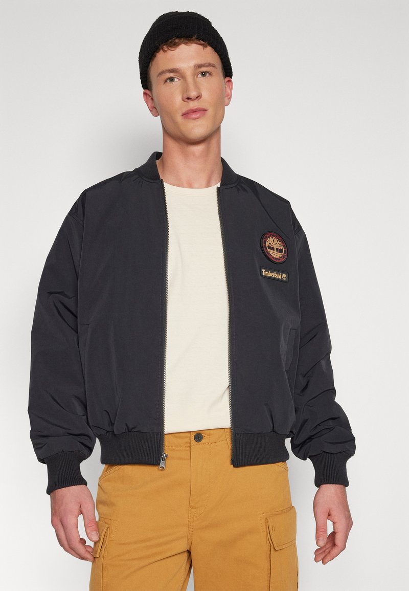 Młody mężczyzna w czarnej czapce beanie, czarnej kurtce bomberce zapinanej na zamek z logo Timberland, białej koszuli i musztardowych spodniach cargo na jednolitym tle.