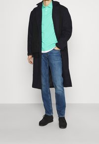 Man som bär en lång svart kappa, mintgrön polo skjorta, blå jeans, svarta skor och svart scarf, stående mot en vit bakgrund.