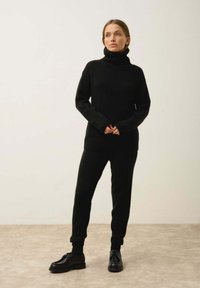 Mustat neulotut korkeakauluksiset neuleet ja siihen sopivat istuvat housut ribatulla hihansuulla, yhdistettynä mustiin nauhakengiin neutraalilla taustalla.