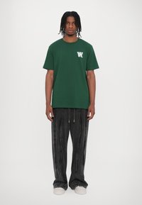 Dark green cotton t-shirt with short sleeves, featuring a white logo on the left chest, paired with loose black denim pants and white shoes.