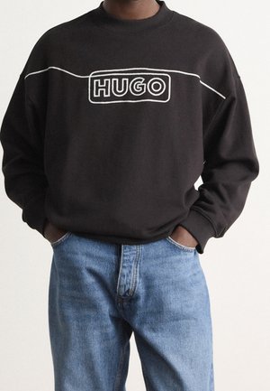 Osoba ubrana w czarną bluzę z białym logo "HUGO" i jasnoniebieskie dżinsy, ręce wsunięte w przednie kieszenie, stoi na jednolitym tle.