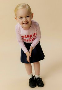 Camicia a maniche lunghe rosa chiaro con la scritta "SPARKLE BRIGHT" in rosso, abbinata a una gonna blu navy. Scarpe nere e calzini bianchi completano l'outfit.