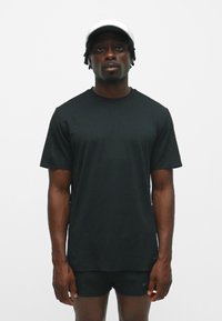 T-shirt noir à manches courtes en tissu lisse, associé à un short noir. Le modèle porte une casquette blanche et se tient devant un fond uni.