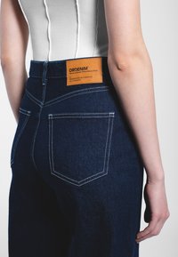 Tmavě modré džíny z denimu s kontrastním bílým šitím, štítkem na zadním pásu a dvěma zadními kapsami obdélníkového tvaru.