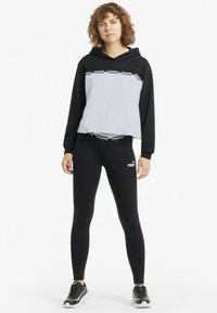 Sudadera con capucha en blanco y negro de diseño cropped, con una banda de logo en el pecho. Leggings negros y zapatillas completan el atuendo.